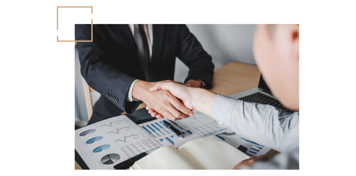 Zwei Personen in Businesskleidung geben sich über einem Tisch mit Diagrammen und einem Notizbuch die Hand.