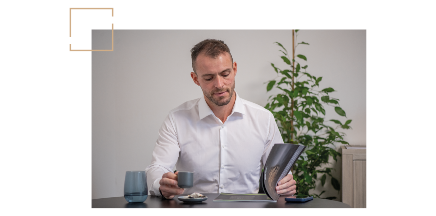 Mann in weißem Hemd sitzt an Tisch, hält Espressotasse und aufgeschlagenes Magazin, im Hintergrund Pflanze.