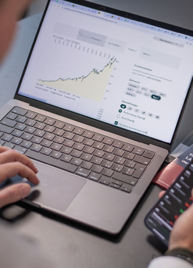 Person sitzt an einem Tisch mit Laptop, auf dem ein Diagramm zu sehen ist, und hält ein Smartphone in der Hand. Auf dem Tisch liegen Magazine, eine Tasse auf Untertasse und ein Glas.