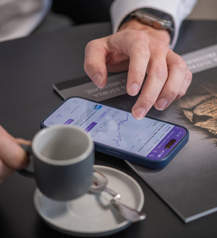 Hand zeigt auf Smartphone mit Finanzdiagramm, daneben Tasse mit Untertasse und Löffel auf Tisch.